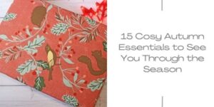 A patterned autumn-themed gift box with leaves, berries, a bird, and a squirrel sits on a white wooden surface next to text reading, 15 Cosy Autumn Essentials to See You Through the Season—perfect for your cosy night in essentials.
