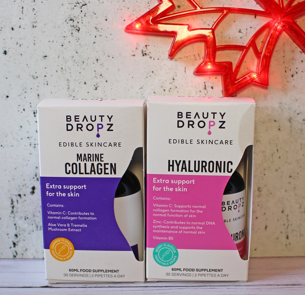 Two boxes of Beauty Dropz edible skincare supplements, labelled Marine Collagen and Hyaluronic, are displayed side by side on a white surface with a red maple leaf light decoration in the background.