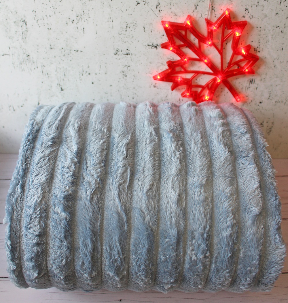 A folded, ribbed, light grey faux fur blanket sits on a wooden surface with a red, illuminated maple leaf decoration on the white wall behind it.