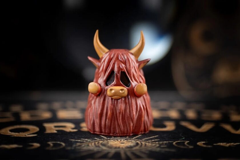 A close-up of a small, hand-crafted figurine resembling a highland cow with long brown fur, beige horns, a wide pink nose, and black hollow eyes, positioned on a dark, patterned surface.