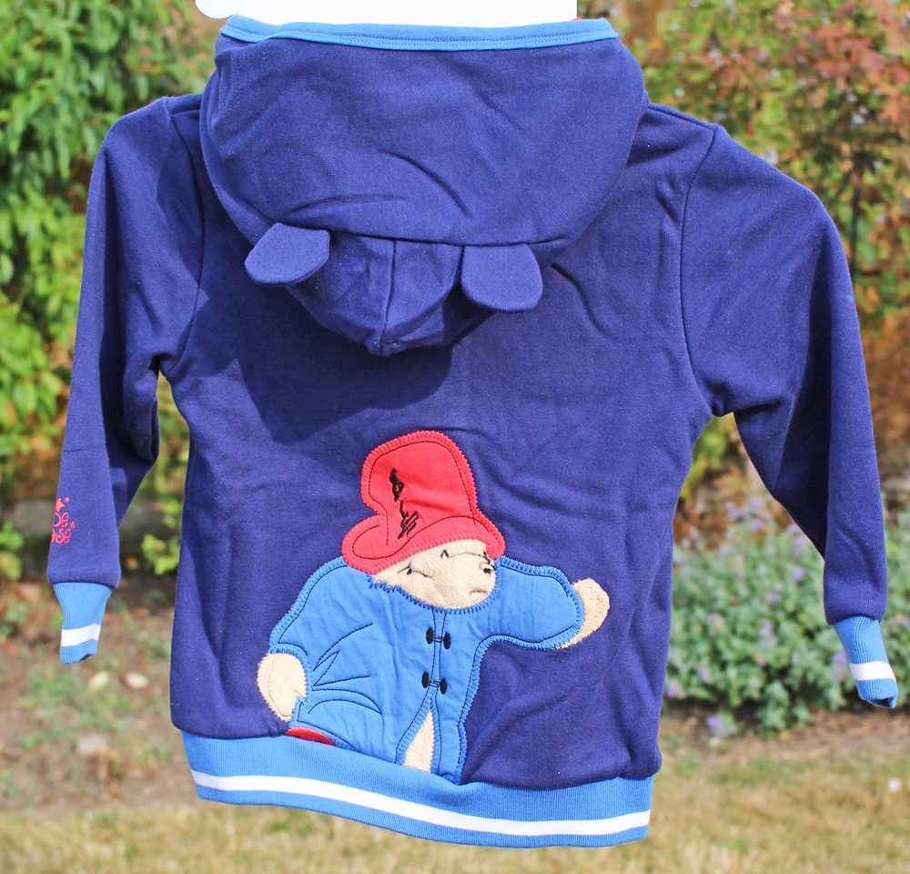 A navy blue children's hoodie with a hood featuring small ears and an embroidered Paddington Bear wearing a red hat and blue coat on the back, displayed outdoors with greenery in the background.