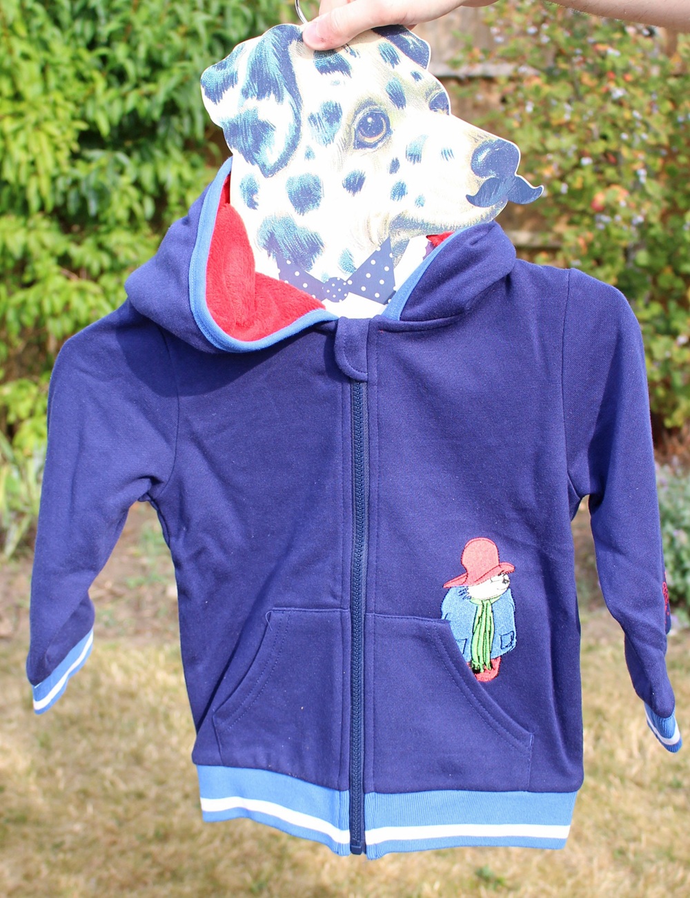 A person holds up a blue hooded children's jacket with Paddington Bear on the pocket and a red-lined hood. In place of a head, there is a cut-out of a spotted dog's face held above the collar. The background is a garden.