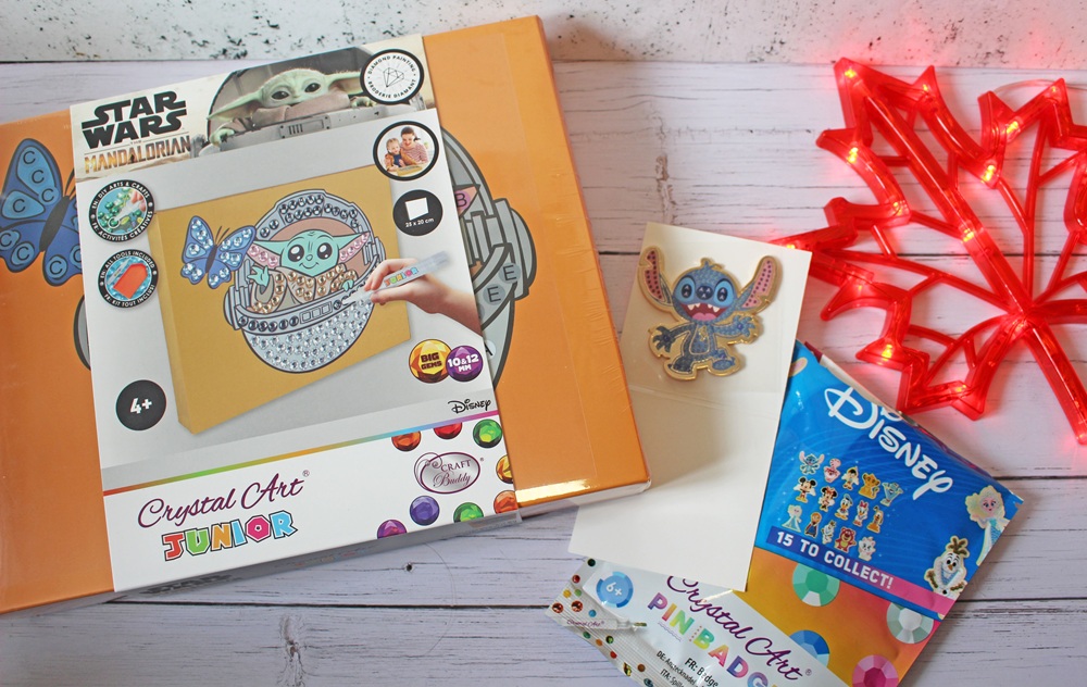 A Star Wars Mandalorian-themed Crystal Art Junior kit is shown, featuring a Baby Yoda design in progress. Nearby are a Disney collectable pin pack, a decorative badge, a crystal art guide, and a red maple leaf light.