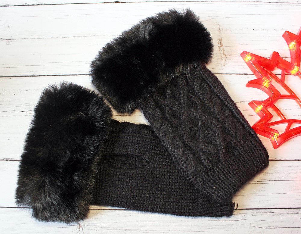 A pair of knitted black mittens with thick, soft faux fur cuffs are laid out on a white wooden surface next to a glowing red star-shaped light decoration.