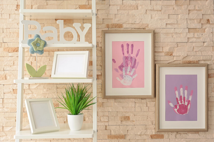 A white shelf with empty frames, a potted plant, and a “Baby” sign stands next to two framed handprint artworks in pink and purple on a beige brick wall.