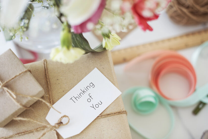 A gift wrapped in brown paper and string with a tag that says Thinking of You, surrounded by pastel ribbons, a ruler, and a blurred vase of flowers in the background.