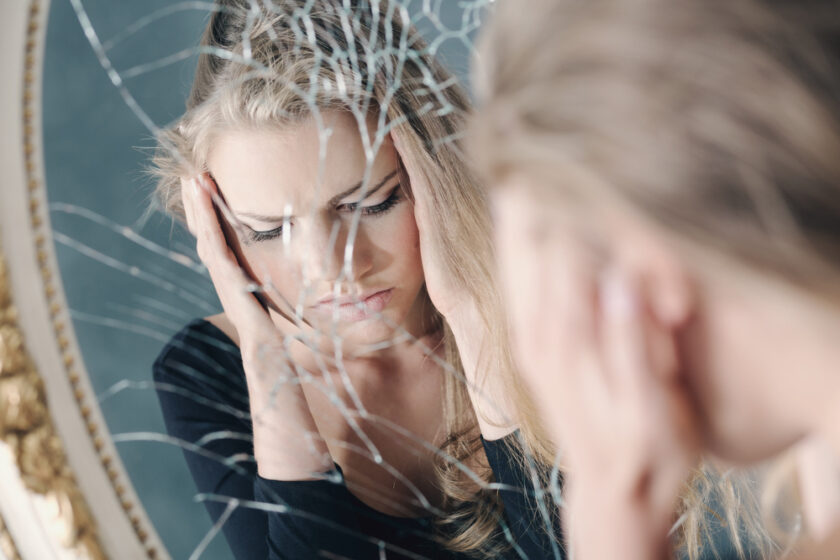 A woman holds her head in her hands and looks distressed whilst staring into a cracked mirror, her reflection fractured by the broken glass.