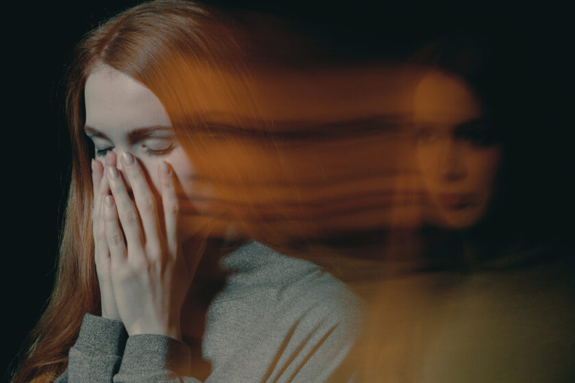 A woman with long red hair covers her face with her hands, eyes closed, looking distressed. A blurred, translucent duplicate of her face appears to the right, suggesting emotions such as anxiety or inner conflict.