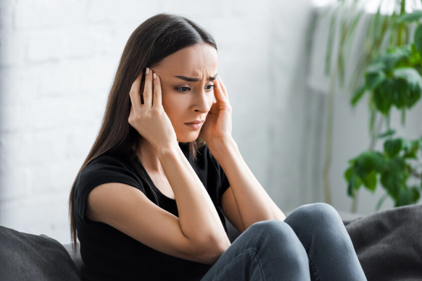 A woman with long dark hair sits indoors, holding her head with both hands and looking distressed or worried. She is wearing a black t-shirt and appears to be deep in thought. Green plants are in the background.