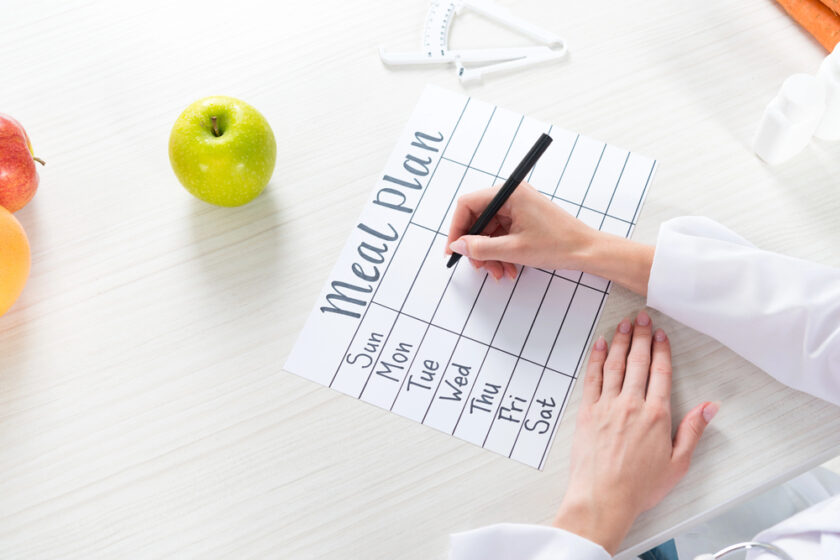 A person writes on a blank weekly meal plan chart at a white table with an apple, orange, and measuring tape nearby.