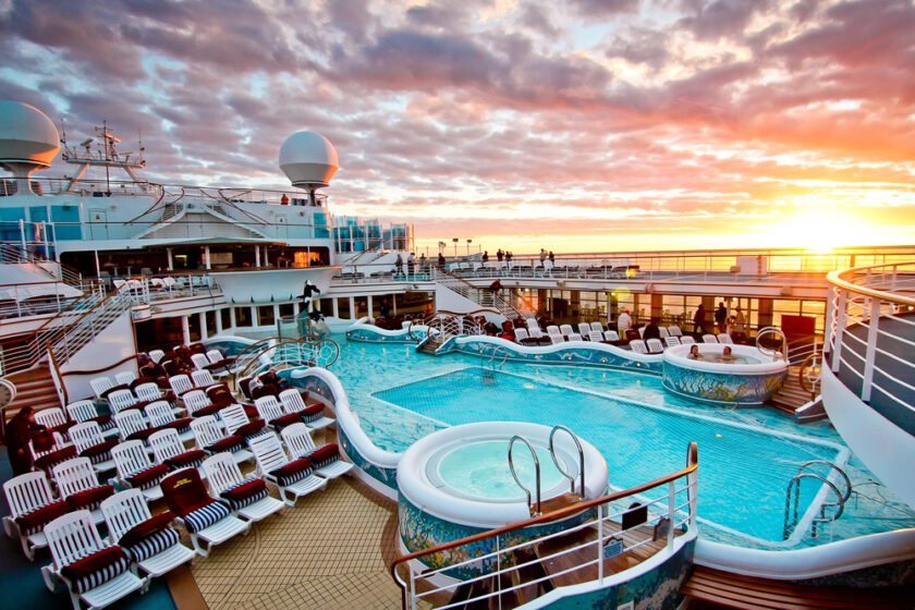 A cruise ship deck features empty sun loungers, a large swimming pool, and hot tubs under a vibrant sunset sky, with the sea visible in the background.