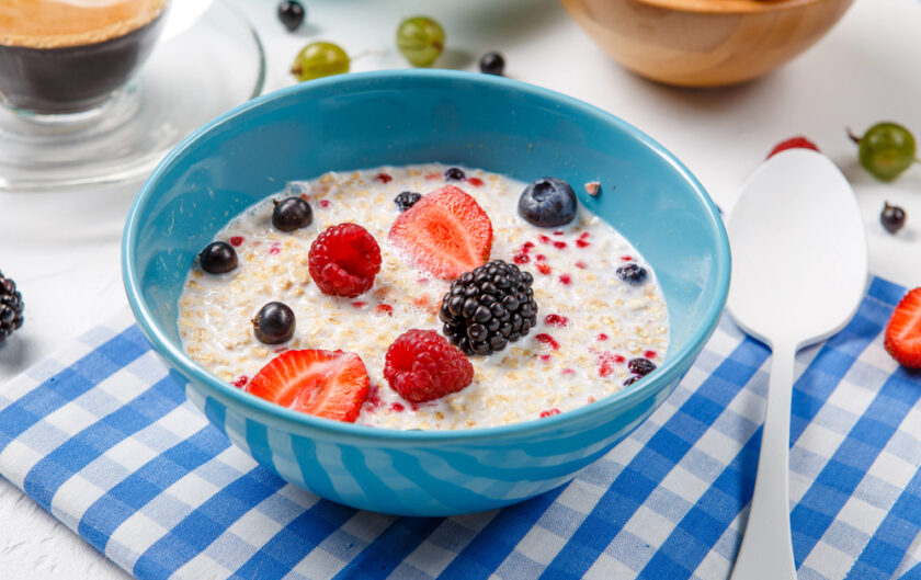 A blue bowl of porridge with milk, topped with strawberries, raspberries, blackberries, and blueberries, sits on a blue chequered serviette next to a white spoon.