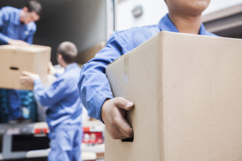 Three people in blue uniforms are loading or unloading cardboard boxes from a lorry. The focus is on a person in the foreground holding a box, with two others handling boxes near the lorry in the background.