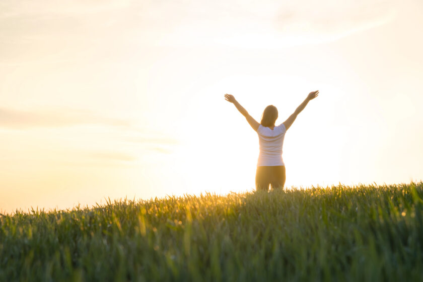A person kneels in a grassy field at sunrise or sunset, facing the sun with arms raised in celebration or gratitude. The sky is bright and the scene evokes a sense of peace and freedom.