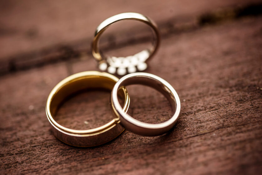 Three rings on a wooden surface: a gold band, a silver band, and a silver ring with small gemstones. The rings are arranged closely together, with soft focus highlighting their textures.