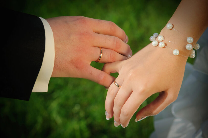 Close-up of a couple’s hands wearing wedding rings, with one hand in a black suit sleeve and the other in a white dress, featuring a pearl bracelet, set against a blurred green background.