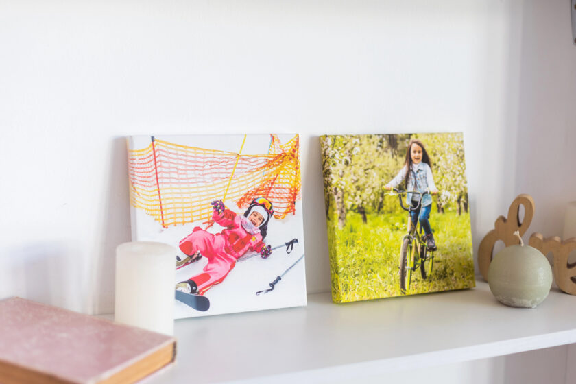 Two framed photos sit on a white shelf: one shows a child skiing in pink snow gear, and the other shows a child riding a bicycle in a green, outdoor setting. A candle, book, and decorative items are nearby.