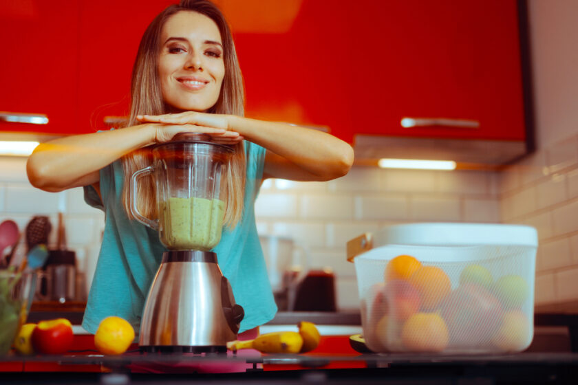A smiling woman in a teal shirt leans on a blender filled with green smoothie in a modern kitchen, surrounded by fruit and a basket of apples on the worktop.