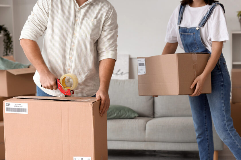 Two people are preparing to move; one is sealing a cardboard box with packing tape, while the other holds another sealed box. Both are standing in a lounge with more boxes and a sofa in the background.