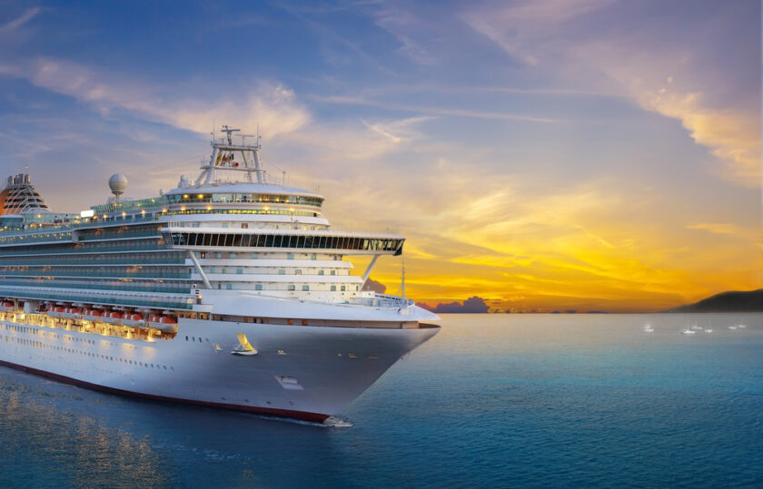 A large cruise ship sails on calm blue water at sunset, with a colourful sky and small boats visible in the distance.