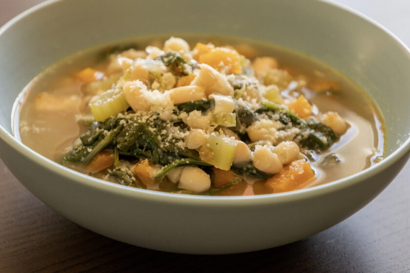 A bowl of vegetable soup containing white beans, chopped carrots, celery, leafy greens, and topped with grated cheese.