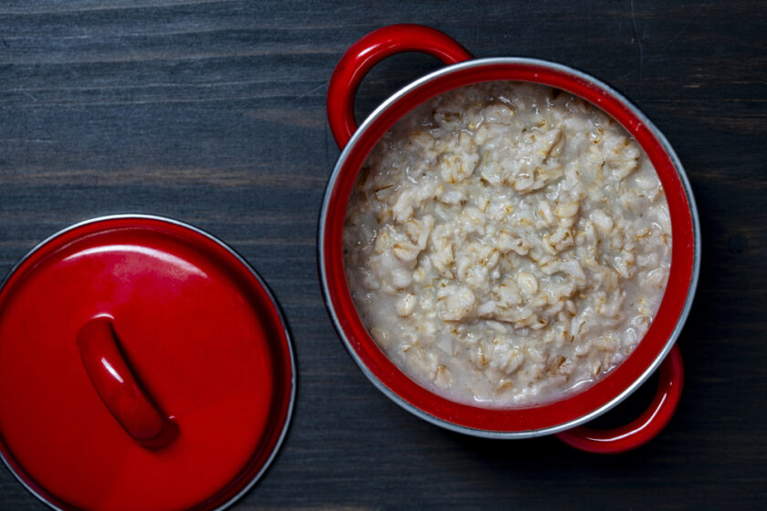 A red saucepan filled with cooked porridge sits on a dark wooden surface, with its matching red lid placed beside it.