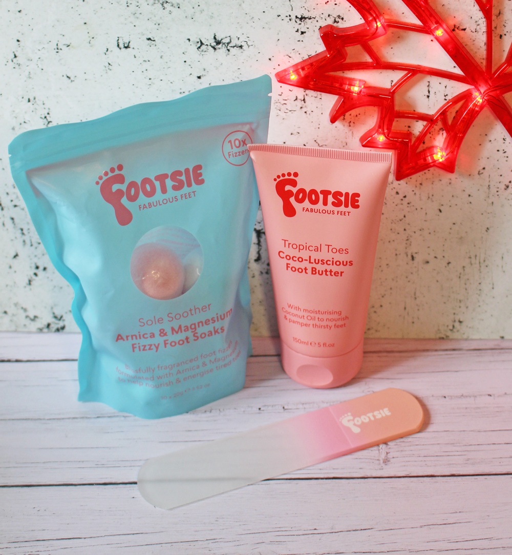 A pastel blue bag of Footsie Arnica & Magnesium Fizzy Foot Soaks, a pink tube of Footsie Tropical Tees Foot Butter, and a Footsie-branded foot file, displayed on a white wooden surface with a red maple leaf light.
