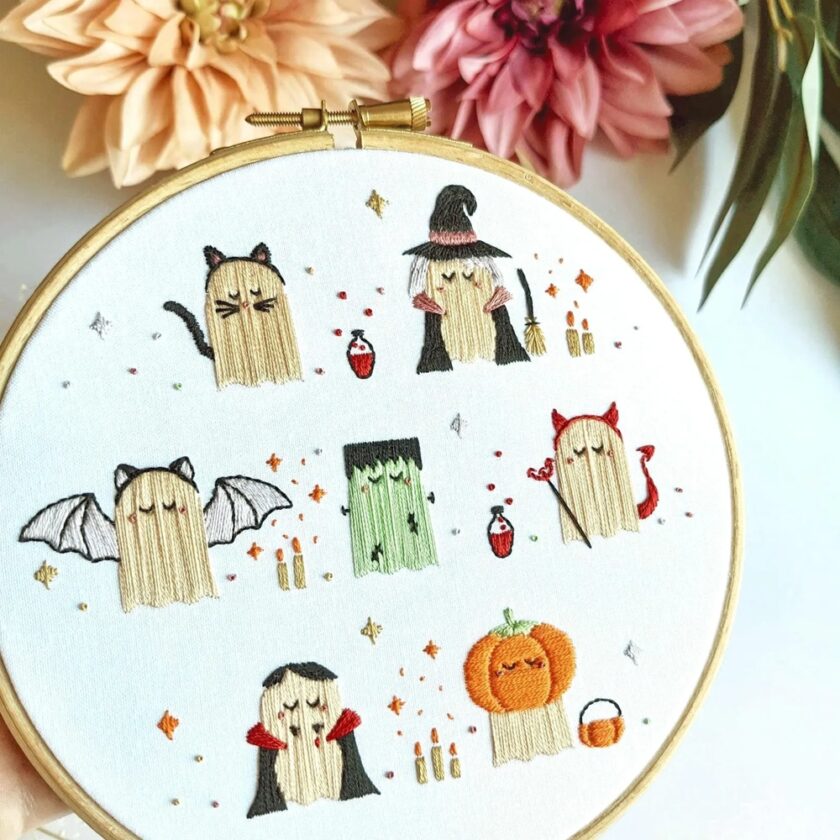 An embroidery hoop features cute ghost figures dressed in Hallowe'en costumes, including a cat, witch, devil, bat, Frankenstein, Dracula, and a pumpkin, with colourful stars and potions around them. Pink flowers are in the background.