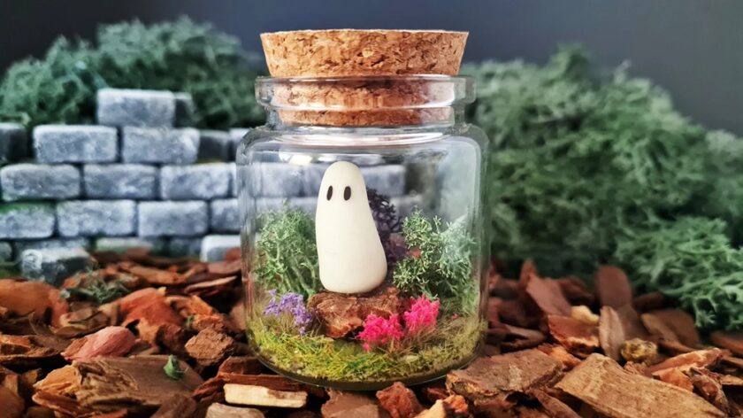A small glass jar with a cork lid contains a cute ghost figurine surrounded by colourful moss and tiny plants, placed on bark chips with stone blocks and greenery in the blurred background.