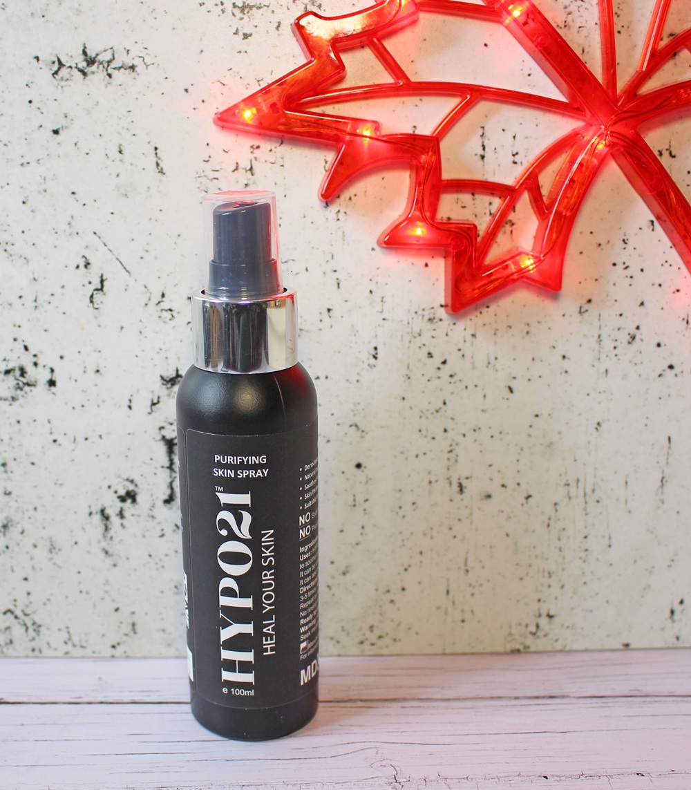 A black bottle of HYPO21 Purifying Skin Spray stands on a white wooden surface beside a red, illuminated maple leaf decoration mounted on a light-coloured, textured wall.