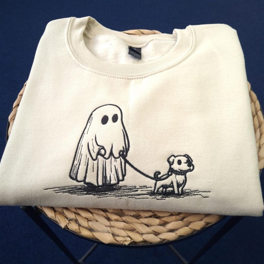 A folded cream-coloured sweatshirt with a simple black line drawing of a ghost figure holding a lead and walking a small dog, displayed on a woven stool against a blue background.