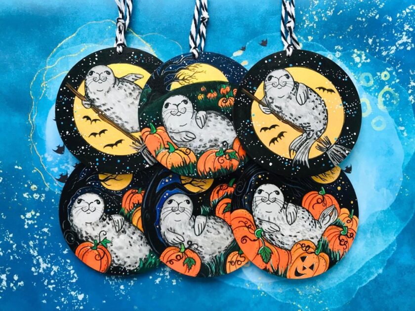 Round baubles with illustrated grey seals lounging among pumpkins and jack-o'-lanterns under starry night skies with full moons. Bats and ravens fly in the background. The baubles are arranged on a blue surface.