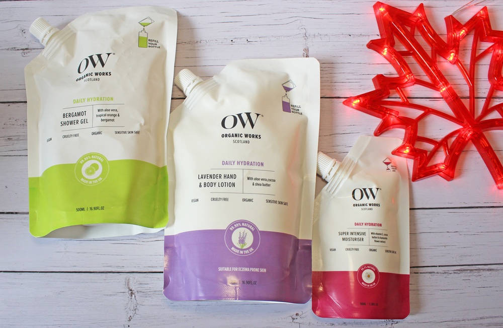Three Organic Works (OW) skincare pouches—a shower gel, hand & body lotion, and moisturiser—are displayed on a white wooden surface beside a red maple leaf-shaped light.