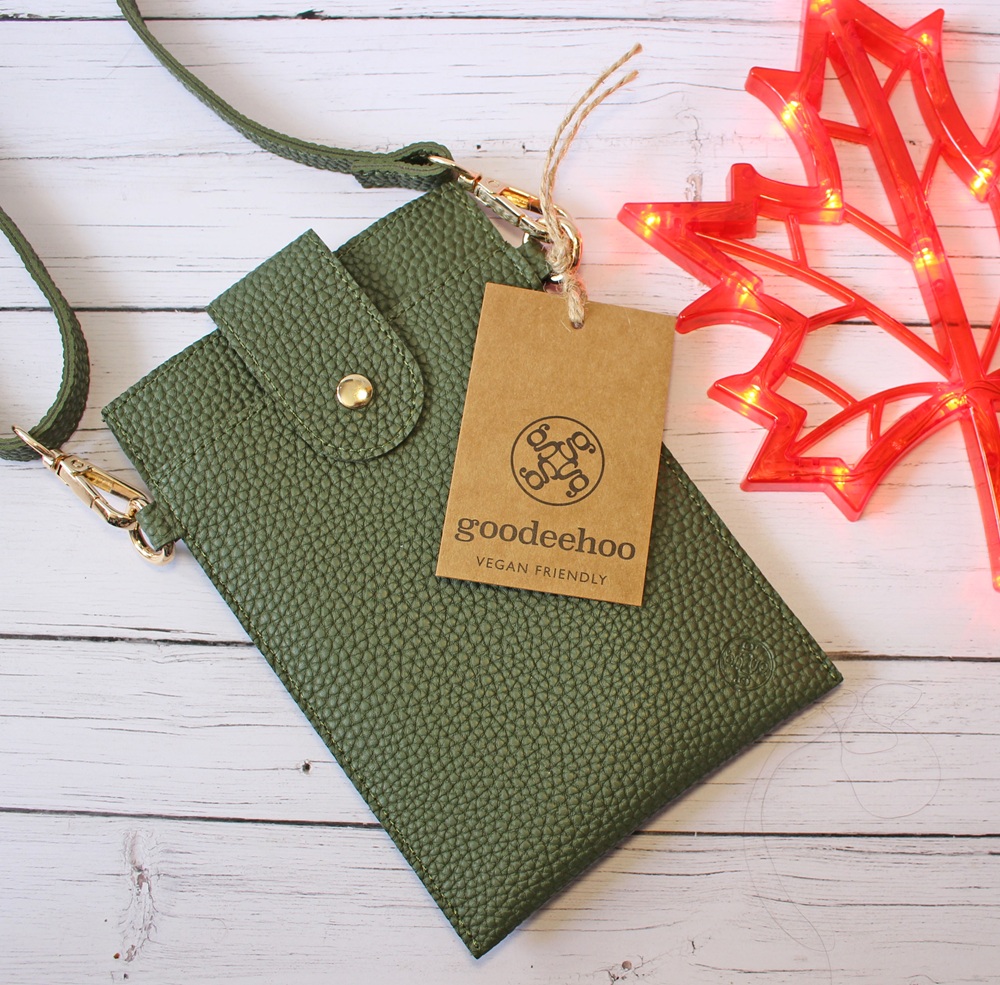 A textured green vegan leather crossbody bag with a popper fastening is displayed on a white wooden surface. A brown tag reads goodeehoo VEGAN FRIENDLY. A red maple leaf-shaped light decorates the corner.