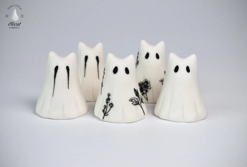 Five small white ceramic ghost figurines stand together, each with simple black eyes; some have black floral or tear-like designs on their bodies. The background is plain and light-coloured.