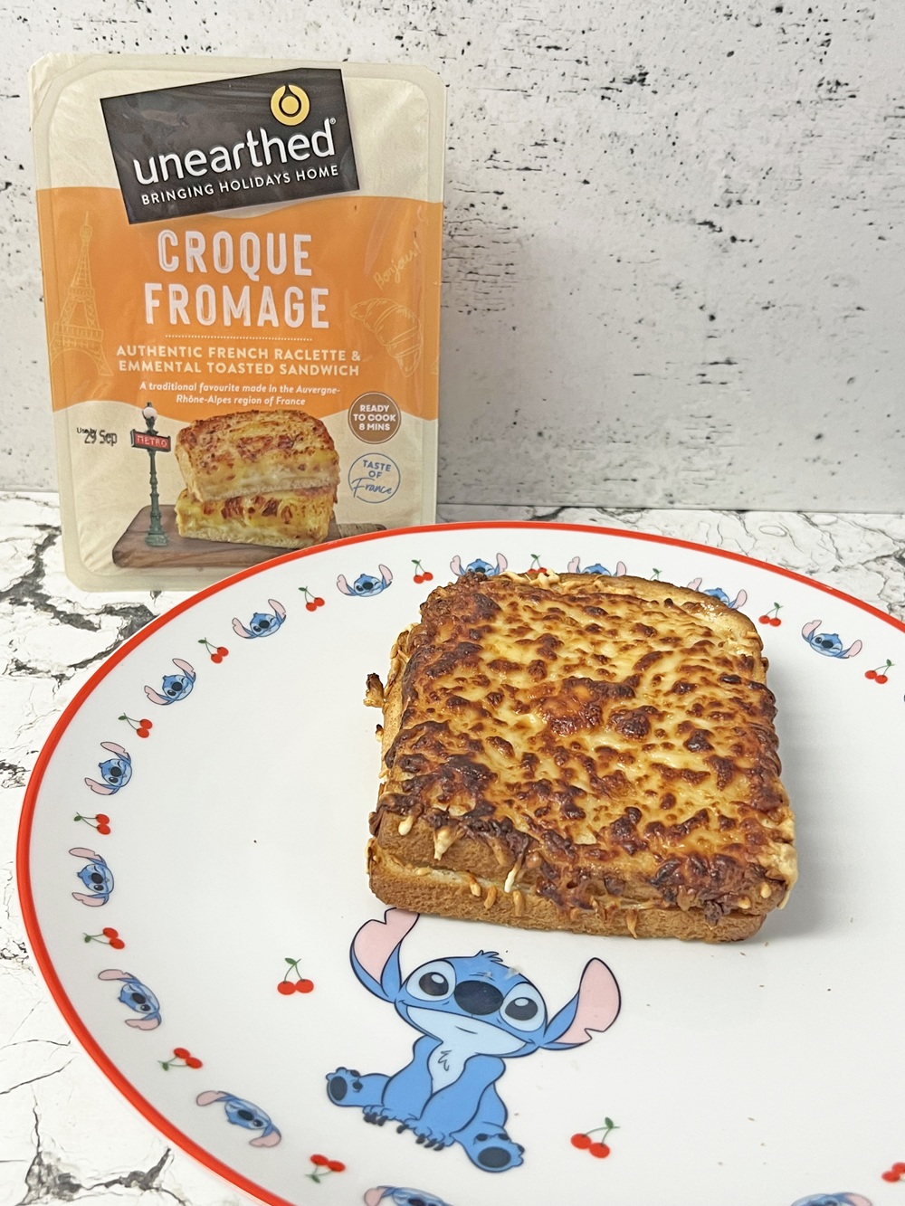 A toasted croque fromage sandwich on a plate with a Stitch cartoon design, in front of a packet labelled unearthed Croque Fromage against a white textured background.