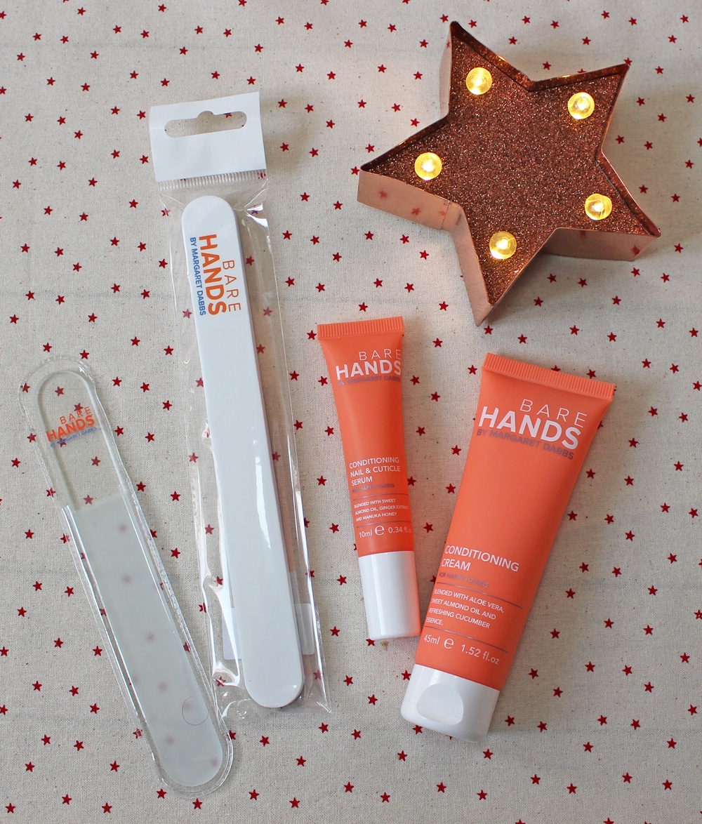 Flat lay of Bare Hands nail care products, including a glass nail file, a nail buffer in packaging, a cuticle cream tube, a conditioning cream tube, and a lit star-shaped decoration on a star-patterned background.