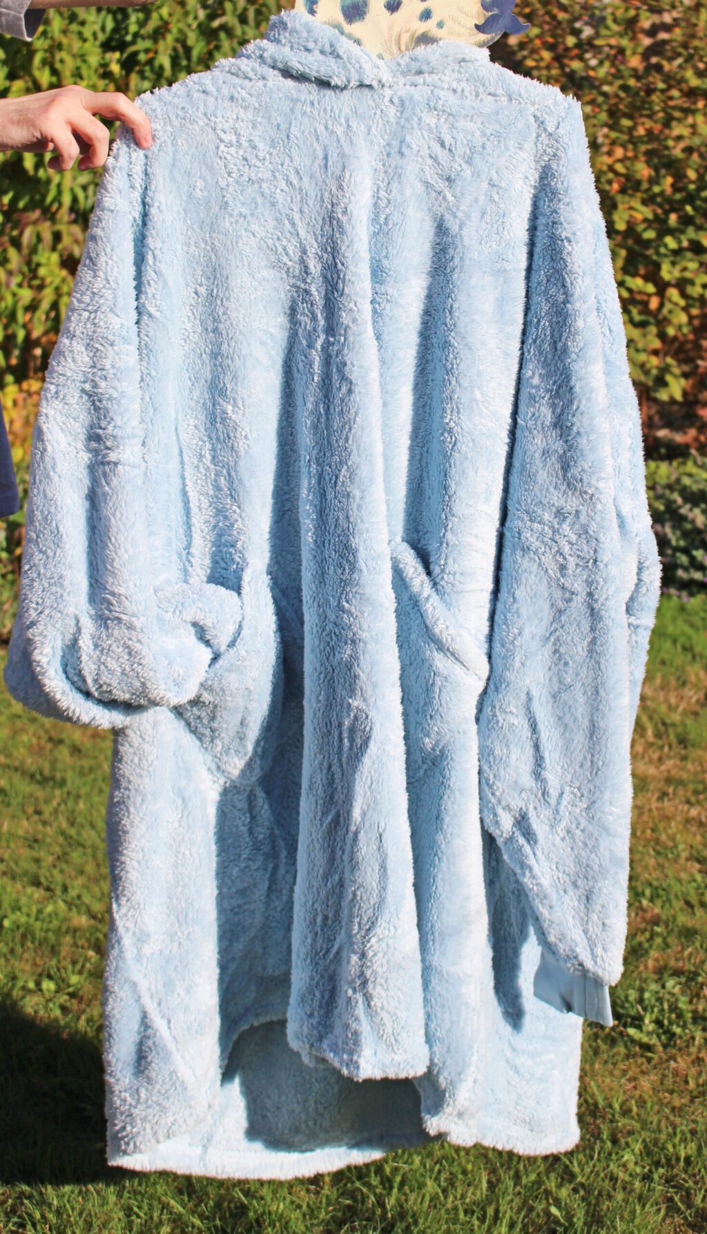 A person holds up a large, light blue, fluffy dressing gown outdoors on a sunny day, with green grass and bushes in the background.