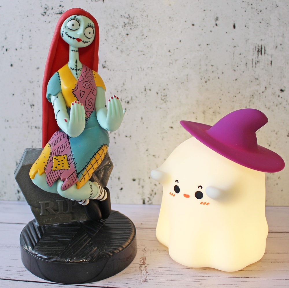 A colourful Sally figurine from The Nightmare Before Christmas sits beside a cute, glowing ghost lamp wearing a purple witch hat on a light wooden surface with a speckled white background.