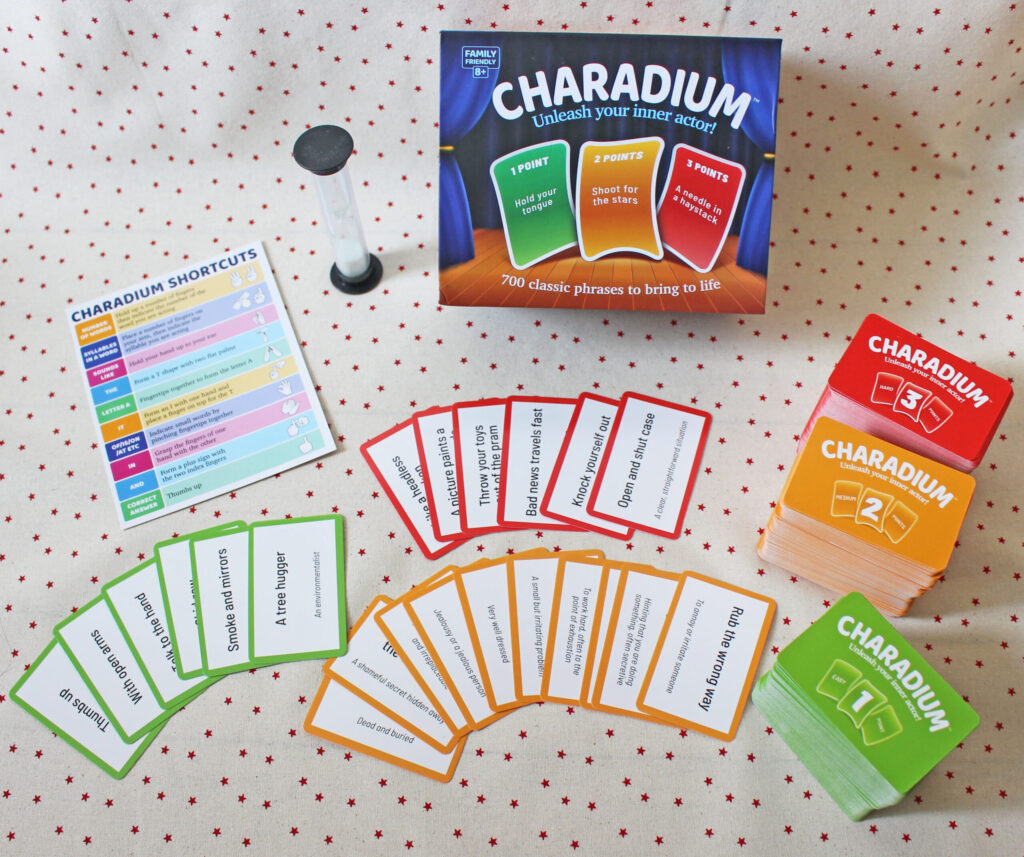 Charadium board game set on a polka dot tablecloth, showing the game box, an hourglass timer, a shortcuts reference card, and various coloured game cards with phrases spread out.