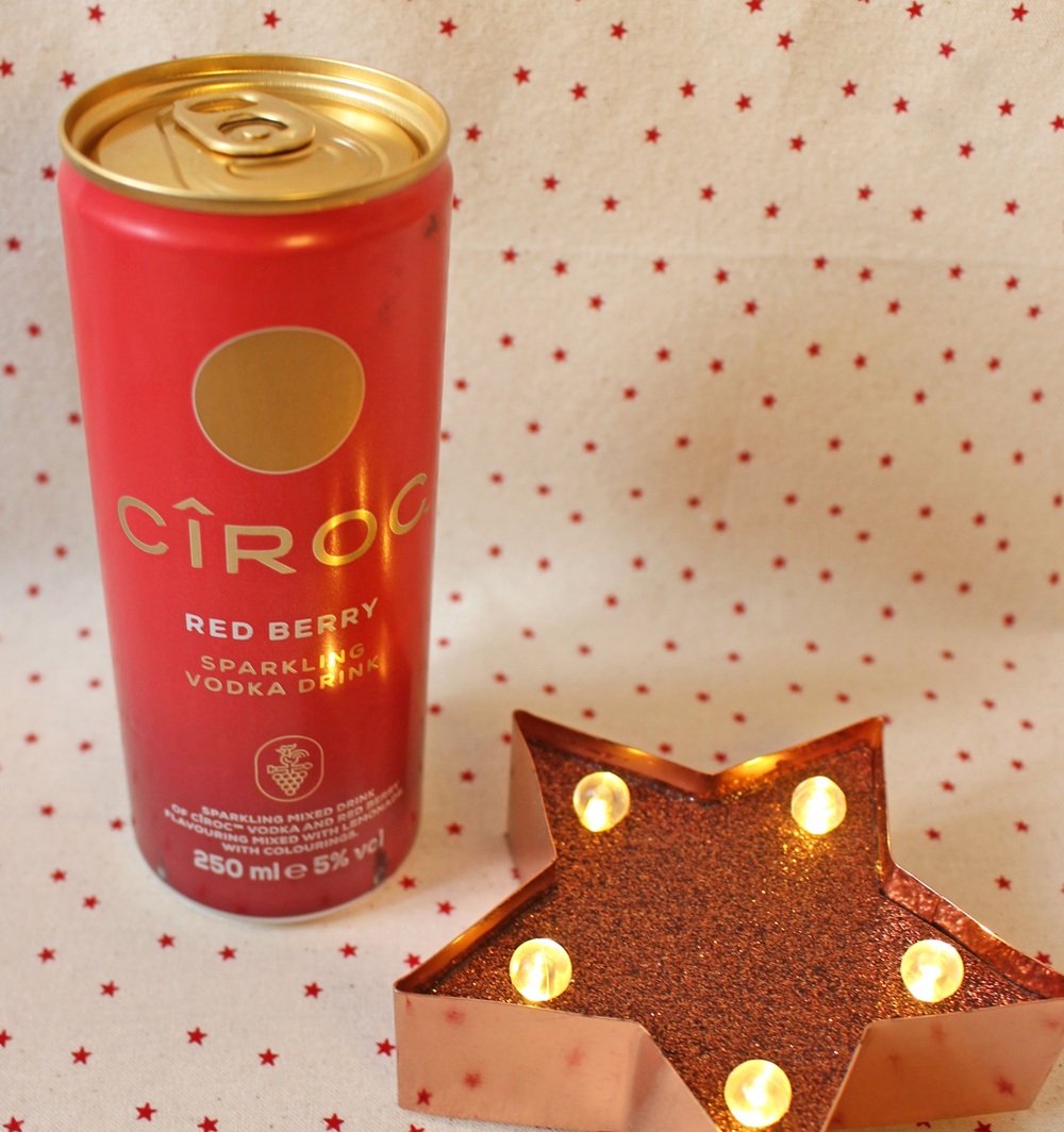 A red can of CÎROC Red Berry Sparkling Vodka Drink stands on a white surface with small red stars, next to a lit star-shaped decoration.