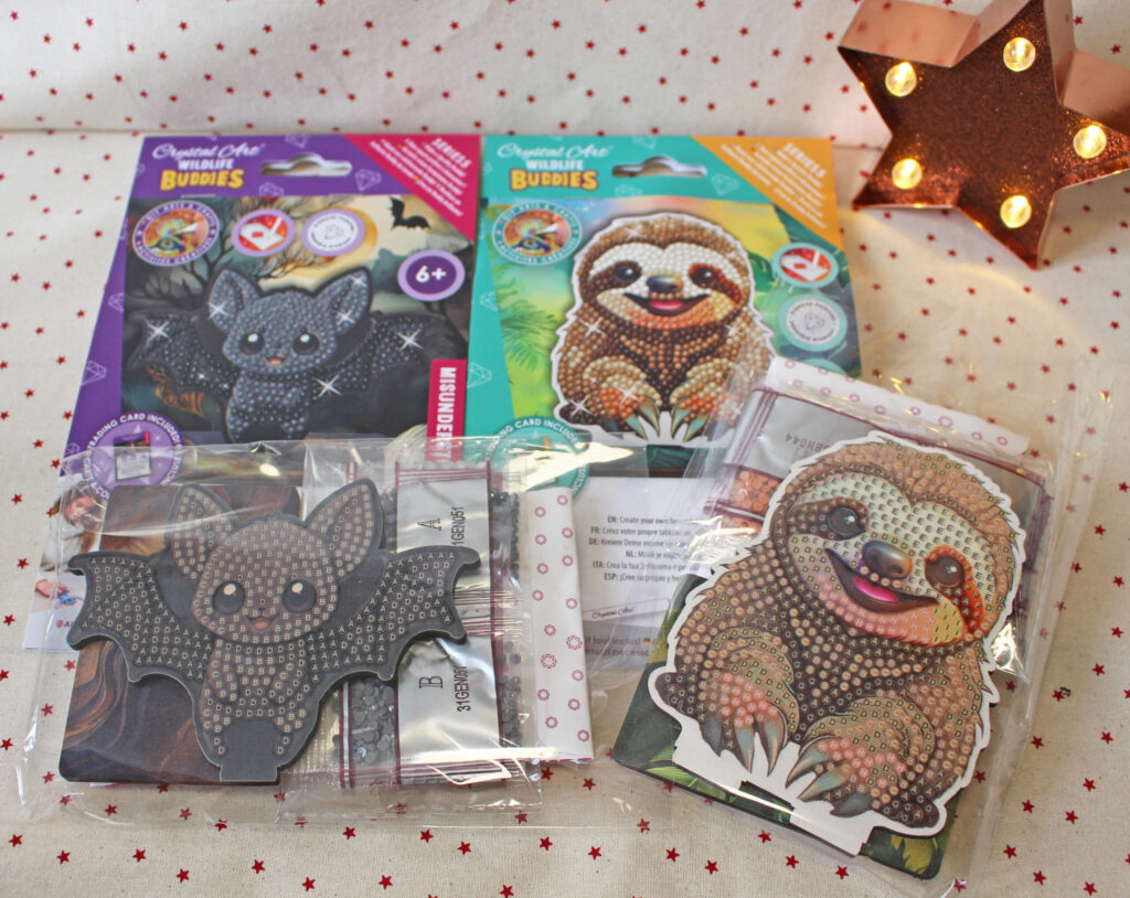 Two diamond art craft kits—one with a bat and one with a sloth—are displayed on a star-patterned cloth. The bat and sloth designs are partially completed with shiny gems. A lit star-shaped lamp is in the background.