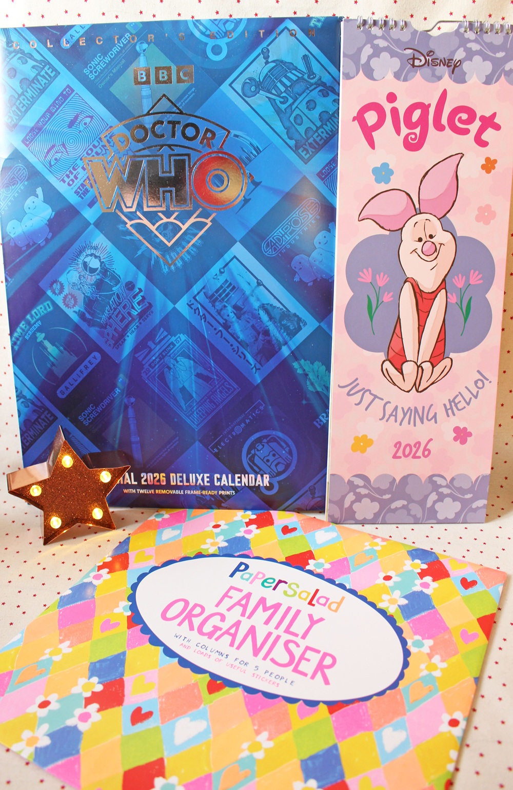 Three 2026 calendars are displayed: a blue Doctor Who calendar, a pink Piglet (Disney) calendar with Just Saying Hello!, and a colourful Papersalad Family Organiser, next to a lit star decoration.