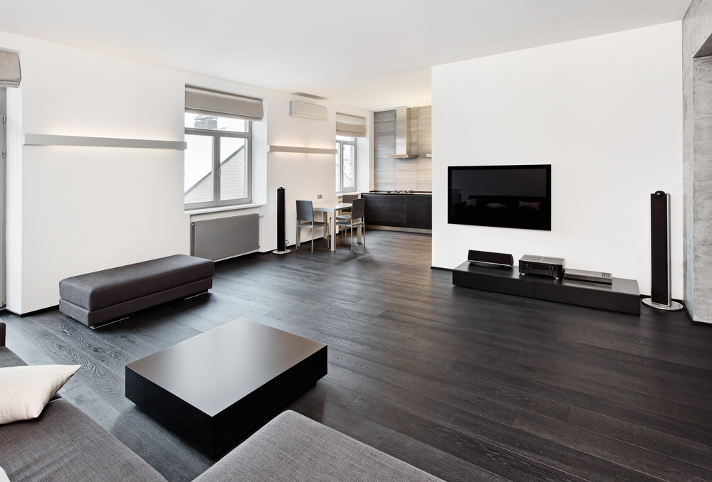 Minimalist living room with dark wood floors, grey furniture, a wall-mounted TV, and a small dining area near the windows. The space is modern, bright, and open with simple decor.