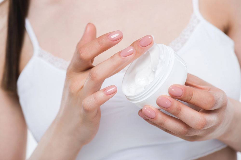 A woman in a white top dips her finger into a white jar of cream, showing neatly manicured nails and smooth skin.