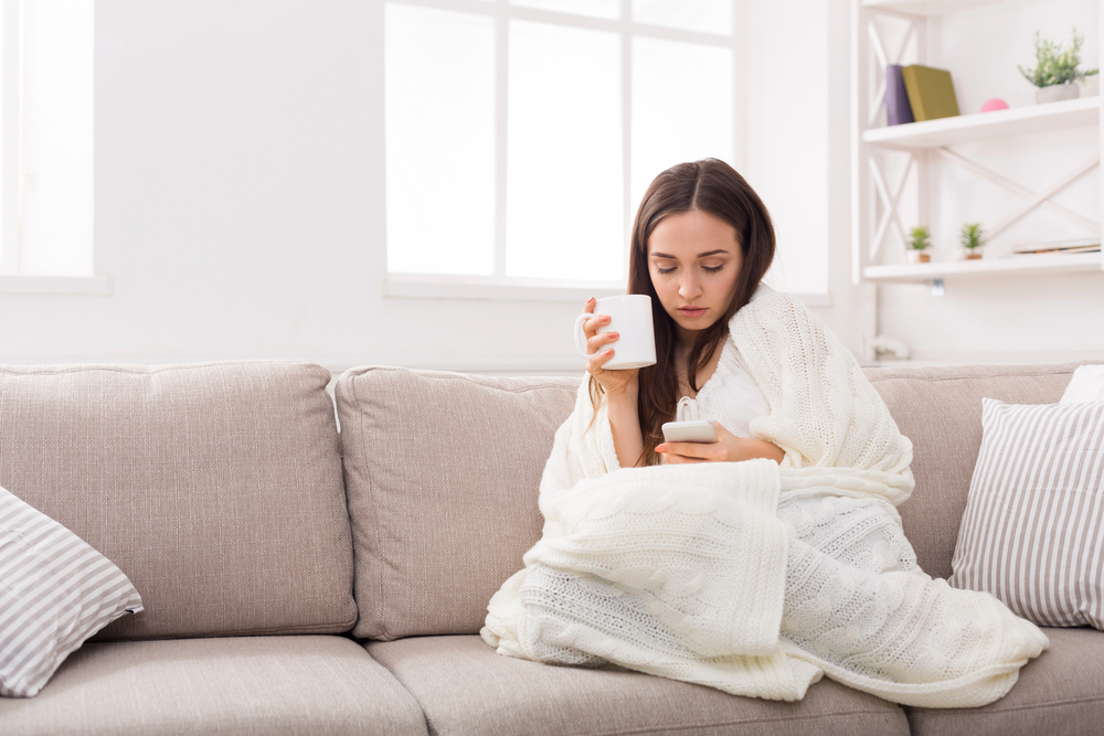 A woman wrapped in a white blanket sits on a beige sofa, holding a white mug and looking at her mobile phone. The room is bright with natural light from a window, and shelves with decor are visible in the background.