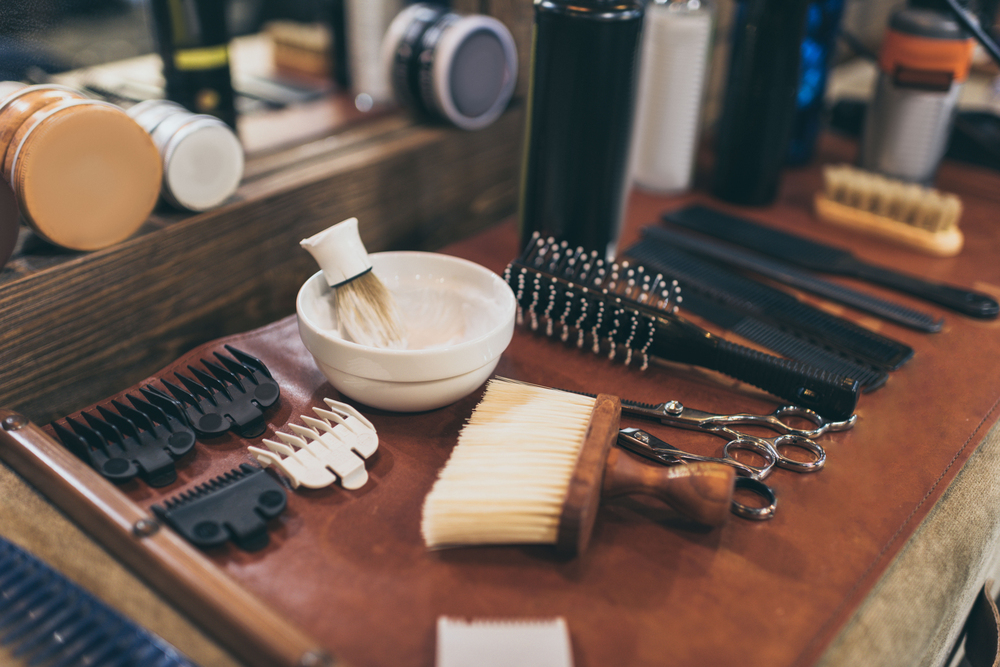 A set of barber tools, including scissors, combs, brushes, clipper guards, a shaving brush in a bowl, and bottles, arranged neatly on a brown mat at a barber’s station.