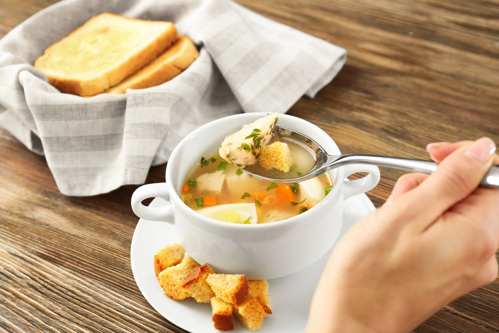 A hand holding a spoon over a bowl of clear soup with vegetables, chicken, and croutons. Toasted bread slices in a cloth-lined basket are in the background on a wooden table.