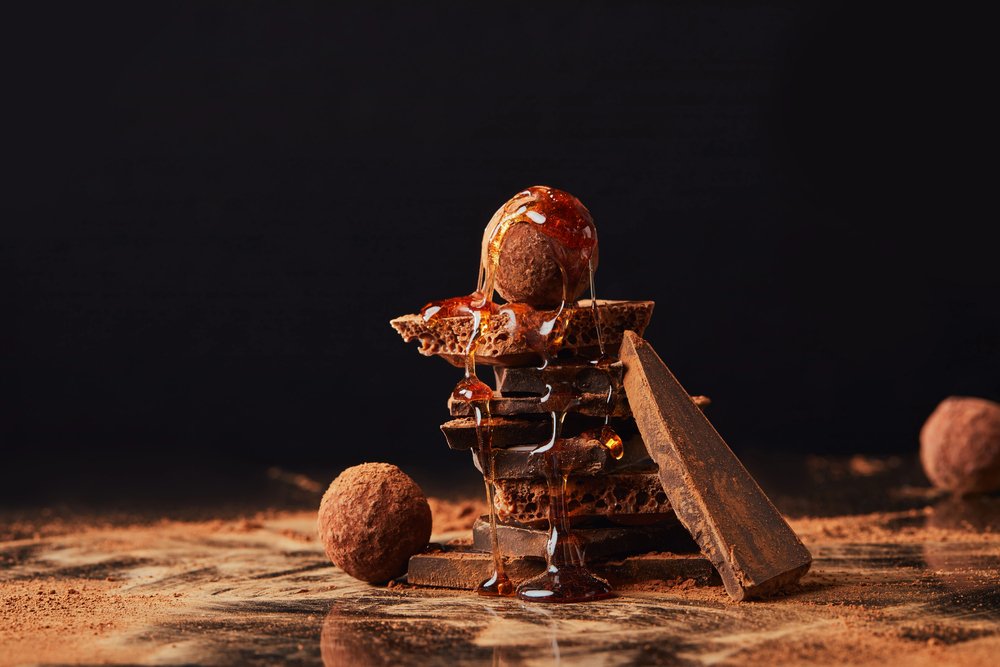 A stack of chocolate pieces topped with a round chocolate truffle and drizzled with syrup, surrounded by cocoa powder and more truffles, set against a dark background.