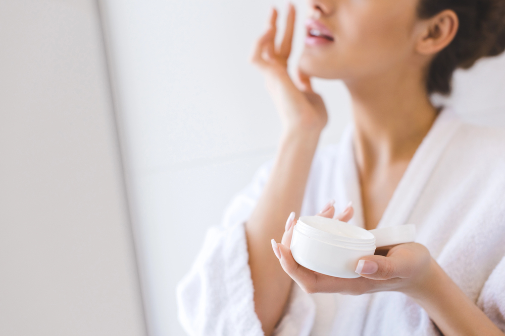 A woman in a white dressing gown applies face cream while holding an open jar of cream, with her face partially out of focus in a bright, softly lit bathroom.